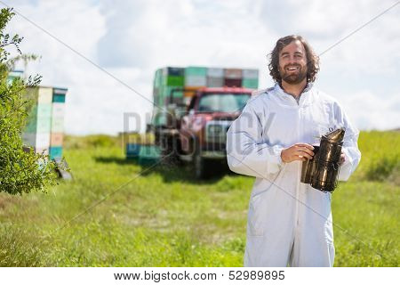 Portrait of happy beekeeper in protective clothing holding smoker while standing at apiary