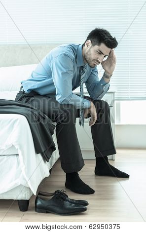 Wincing businessman sitting at edge of bed at home in bedroom