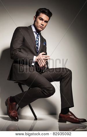 Side view picture of a young elegant business man sitting with one leg in front of the other, looking at the camera.