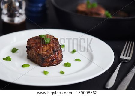 Korean style glazed pork chops on white plate. Sticky citrus caramelized sauce with oranges and mint leaves. Salt, pepper, knife and green mint leaves near pan with chops.