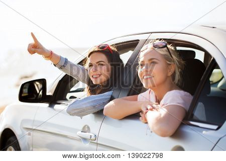 summer vacation, holidays, travel, road trip and people concept - happy teenage girls or young women sitting in car at seaside and pointing finger to something