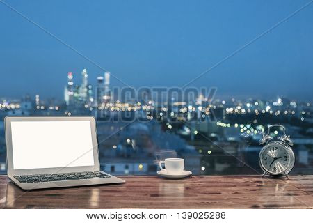 Front view of creative designer desktop with blank white notebook silver alarm clock and coffee cup on blurry night city background. Mock up