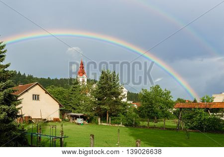 Double rainbow over a church in a cloudy village. Traditional farm with church in a village and double rainbow over woods and trees