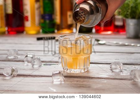 Shaker pours liquid into glass. Orange drink with ice cubes. Fresh grapefruit juice for beverage. Melting ice on wooden table.