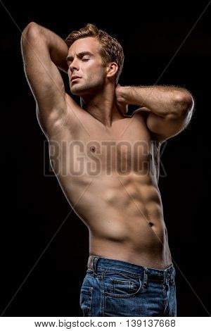 Strong man in blue jeans with naked torso stands with closed eyes on the black background in the studio. He holds his hands on the neck behind the head. Vertical low-key photo.