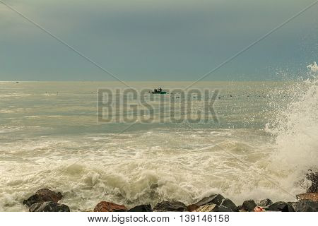 A small Vietnamese fishing boat at sea while waves crash agains the rocky shore