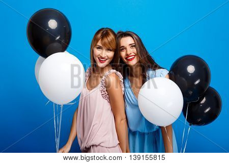Portrait of two young beautiful girls in dresses looking at camera, holding baloons, laughing, smiling, resting at party over blue background.