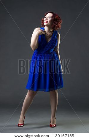 Young beautiful red-haired caucasian woman in blue dress posing in studio on gray background professional makeup and hairstyle full-length portrait
