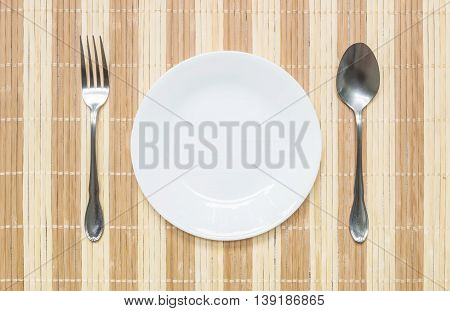 Closeup white ceramic dish with stainless fork and spoon on wood mat textured background on dining table in top view