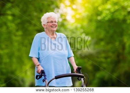 Happy senior handicapped lady with a walking disability enjoying a walk in a sunny park pushing her walker or wheel chair aid and support during retirement concept.