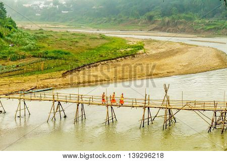 Orange robed buddhist monks walk over a bamboo bridge in Luang Prabang, Laos