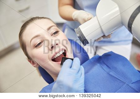 people, medicine, stomatology, technology and health care concept - close up of dentist putting intraoral shield to female patient mouth and assistant directing x-ray machine at dental clinic