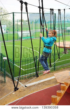 Happy pretty girl climbs on Rope way with safety rope on special playground