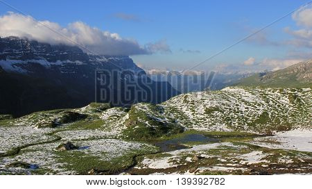 Green meadow with pond valley and mountains. New snow in summer. Swiss Alps.