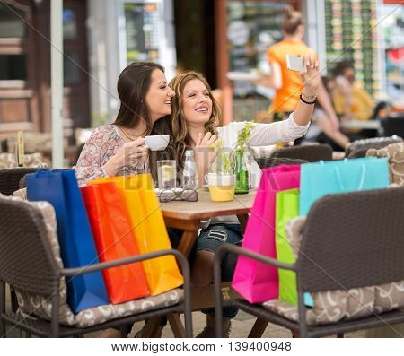 Two smiling female friends driniking coffee and taking selfie after shopping