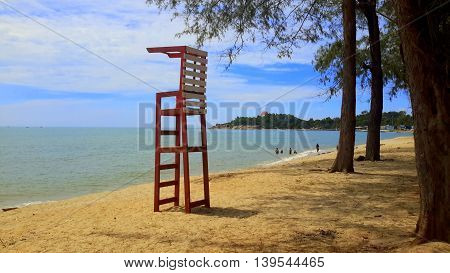 red and white wooden slat lifeguard stand on the beach under pine trees on the right, distant swimmers playing in the sea, peninsula seen in the distant background, Songkhla, Thailand