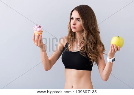 Real temptation. Thoughtful beautiful baffled woman holding cupcake and apple while choosing between them