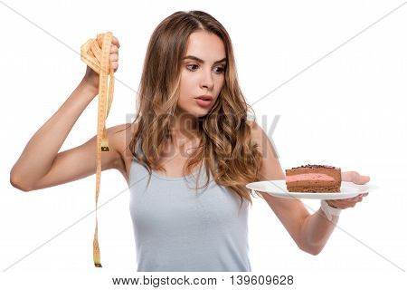 Real dilemma. Pleasant baffled woman holding plate with cake and measure tape and choosing between them while standing isolated on white background