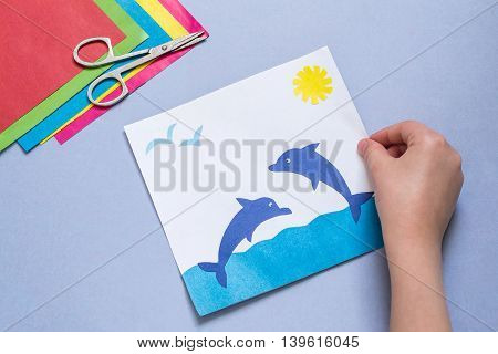 Paper applique is made by the child on a sea theme. The idea for children's creativity an art project made of paper. Sheets of colored paper glue scissors. Children's hand holding a picture