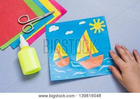 Paper applique is made by the child on a sea theme with sailboats. The idea for children's creativity an art project made of paper. Sheets of colored paper glue scissors. Children's hands holding a picture