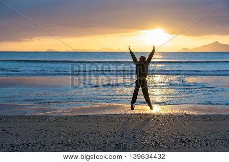 Sunrise glistens over water from horizon and off-shore islands as boy star jumps with happiness on wintry morning at waters edge on beach Waipu Cove New Zealand