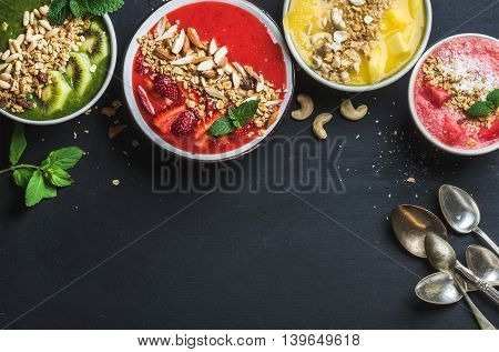 Healthy summer breakfast concept. Colorful fruit smoothie bowls with nuts and oat granola on black background . Yellow red green pink. Top view copy space