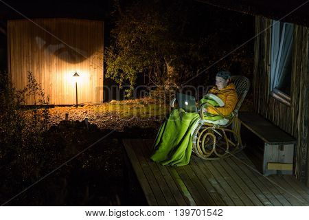 Recreational Village Bungalow and Person sitting in vintage rocker-chair covered by pled working on computer laptop at patio