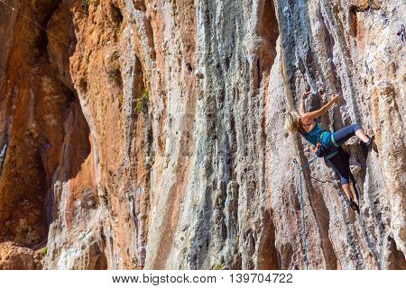 Blond Female Climber ascending Vertical Orange Rocky Wall unusual shape of Mountain Range on background Outdoor Sunshine