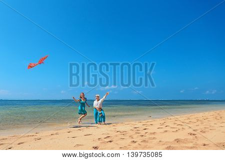 Happy family on beach - grandmother mother and baby girl have fun woman run along sea surf with water splashes launching bird kite. Active parent and people activity on summer holiday with children