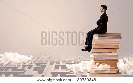 A serious businessman in suit sitting on a pile of giant books in front of a grey wall with clouds, labirynth