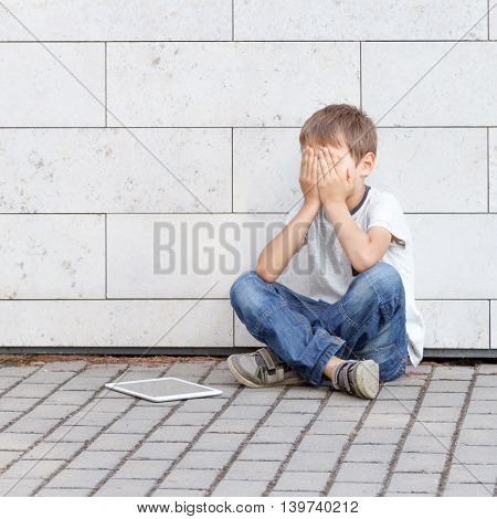 Little boy sad, tired and stressed with tablet computer PC. Kid sitting on the ground, holding his head, close eyes. Casual wear. Outdoor. Education, learning, technology, addiction concept
