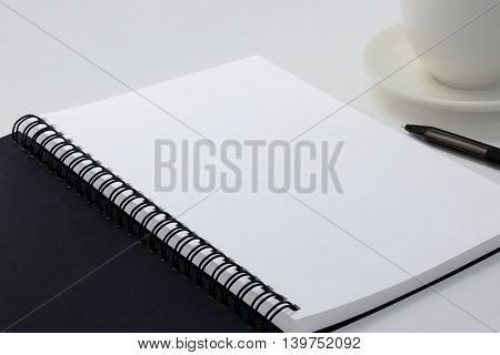 Blank notebook on white desk background with a pen and coffee cup on the right side.