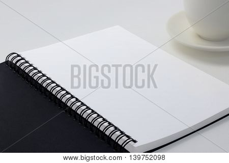 Blank notebook on white desk background and coffee cup on the right side.