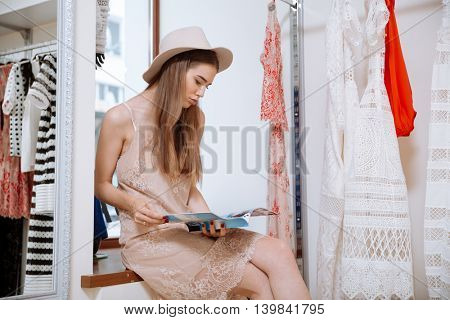 Relaxed attractive young woman in hat sitting and reading magazine in clothing store