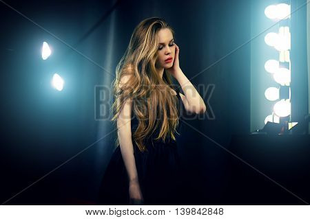 Beautiful sexy young caucasian woman in black dress looking into makeup mirror at herself. Backstage dramatic shot.
