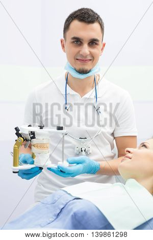 Smiling densist and a female patient in blue shirt and patient bib. Dentist wears white uniform with blue latex gloves and a blue mask, binocular loupes. He holds an articulator with teeth mould. Vertical.