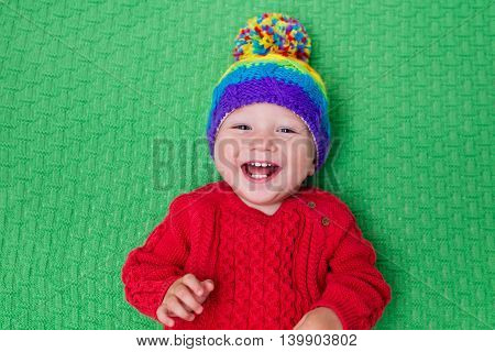 Cute baby in warm wool knitted hat on a red blanket. Autumn and winter clothing for young kids. Colorful knitwear for children. Adorable little boy ready for a walk on a cold fall day.