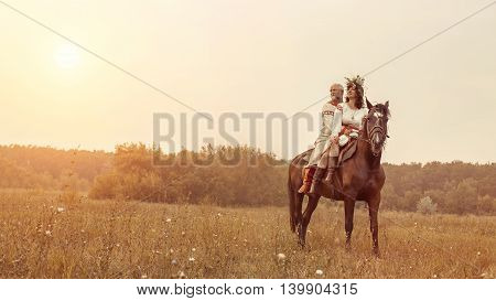 Mature Man And Woman In Ethnic Clothes Are Riding A Horse On The Rural Summer Background.