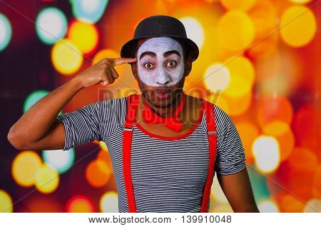 Pantomime man wearing facial paint posing for camera, using hands interacting body language, blurry lights background.