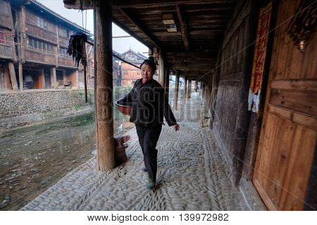 Zhaoxing Dong Village Guizhou Province China - April 9 2010: Asian women rural resident is walking on a paved stone walkway near the river embankment of wooden houses of local farmers.