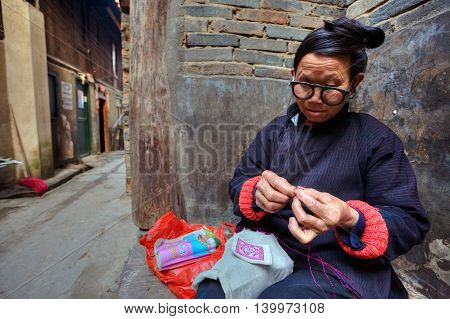 Zhaoxing Dong Village Guizhou Province China - April 9 2010: Mature Asian woman embroiders designs on fabric sitting near the wall of a stone house on a rural street.