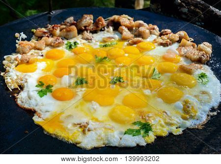 Fried eggs with pieces of pork on a large flat pan cooking outdoors.