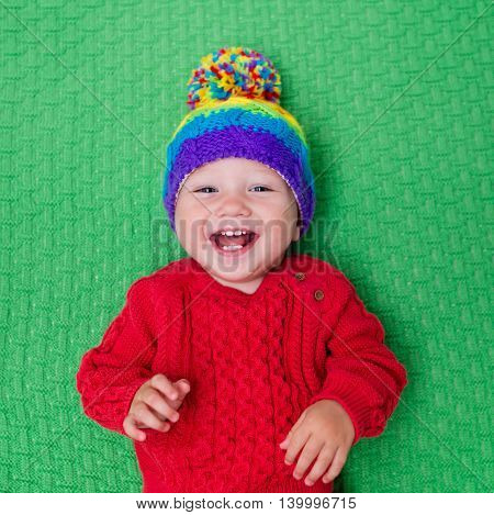 Cute baby in warm wool knitted hat on a red blanket. Autumn and winter clothing for young kids. Colorful knitwear for children. Adorable little boy ready for a walk on a cold fall day.