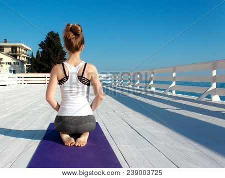 Young Slim Woman In Tight Sportswear Practicing Yoga Outdoors At White Wooden Seafront. Back View Ei