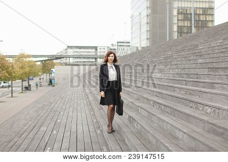 European Businesswoman Standing On Stairs With Bag And High Buildings In Background. Concept Of Prof
