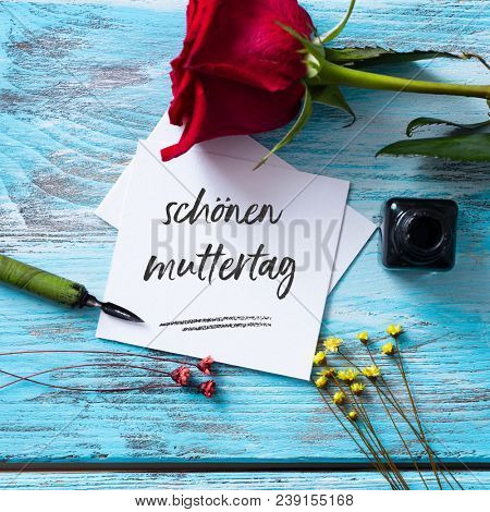 high angle view of a red rose, an ink bottle, a nib pen and a piece of paper with the text schonen muttertag, happy mothers day written in german, on a blue rustic wooden table