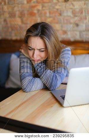 Young Woman Sitting In Cozy Coffee House Near Wooden Table With Glass Cup Of Cappuccino And Using La