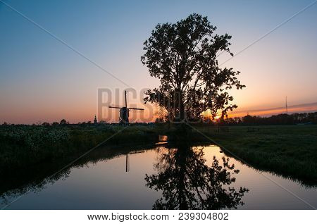 Sunrise In Dutch Village. Old Tree And Traditional Dutch Windmill Reflected In The Calm Water Surfac