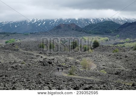 couple hikers on the path to extinct cinder cones under the majestic volcanic cone of Etna Mount that is covered by clouds, Sicily