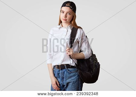 Studio Shot Of Pretty Lovely Schoolgirl Wears Jeand And White Shirt, Carries Rucksack, Stands Agains